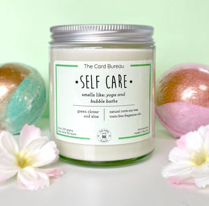 Photo of Self Care Candle, surrounded by bath bombs and flowers against a pale green backdrop