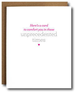 Minimalist greeting card with the message 'Here’s a card to comfort you in these unprecedented times' in pink and gray text, centered on a white background with a small pink heart below. Brown kraft paper envelope partially visible behind the card.