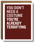 A greeting card with a dark red background and bold white text reading 'YOU DON'T NEED A COSTUME YOU'RE ALREADY TERRIFYING', shown with a brown kraft envelope behind it on a white background.