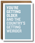 A greeting card with a light blue background and bold white text reading 'YOU'RE GETTING OLDER AND THE COUNTRY'S GETTING WEIRDER,' shown with a brown envelope behind it on a white background.
