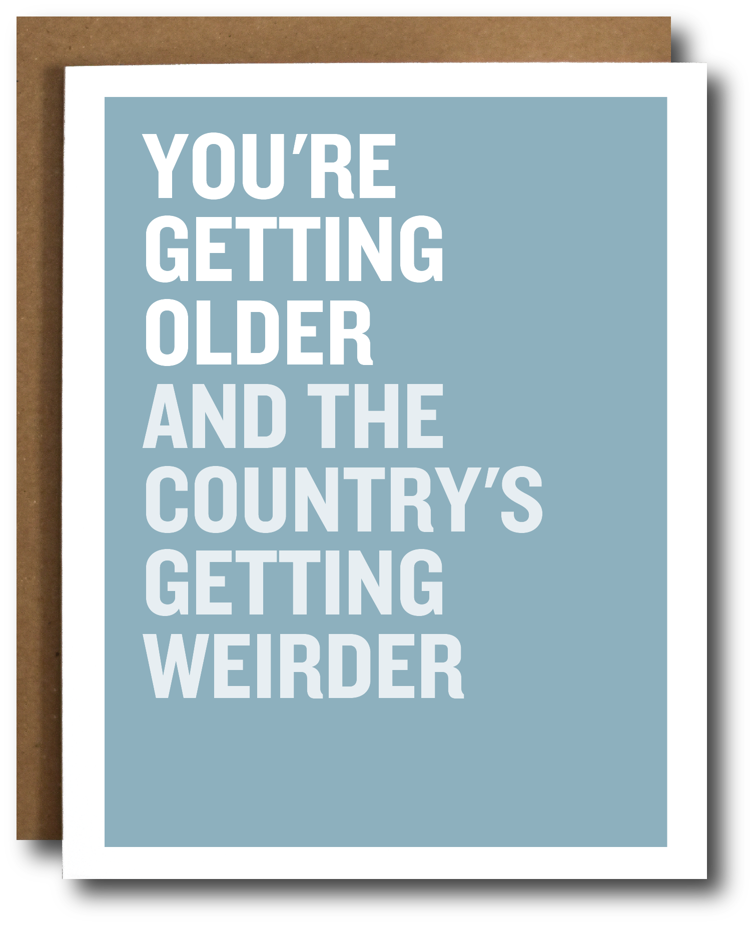 A greeting card with a light blue background and bold white text reading 'YOU'RE GETTING OLDER AND THE COUNTRY'S GETTING WEIRDER,' shown with a brown envelope behind it on a white background.