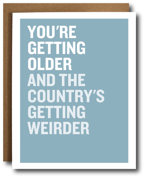 A greeting card with a light blue background and bold white text reading 'YOU'RE GETTING OLDER AND THE COUNTRY'S GETTING WEIRDER,' shown with a brown envelope behind it on a white background.