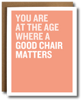 A greeting card with a salmon pink background and bold white text reading 'YOU ARE AT THE AGE WHERE A GOOD CHAIR MATTERS', shown with a brown kraft envelope behind it on a white background.
