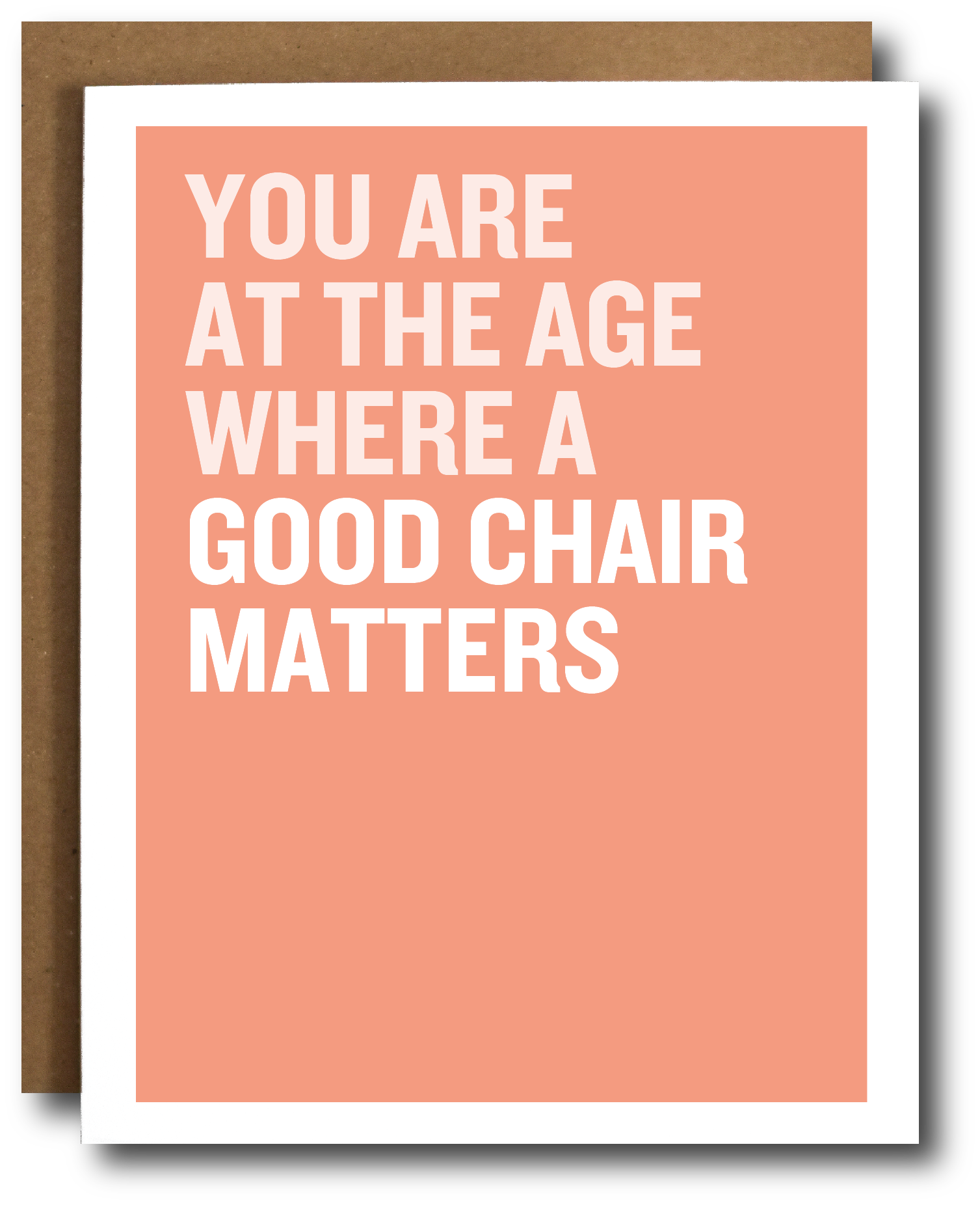 A greeting card with a salmon pink background and bold white text reading 'YOU ARE AT THE AGE WHERE A GOOD CHAIR MATTERS', shown with a brown kraft envelope behind it on a white background.