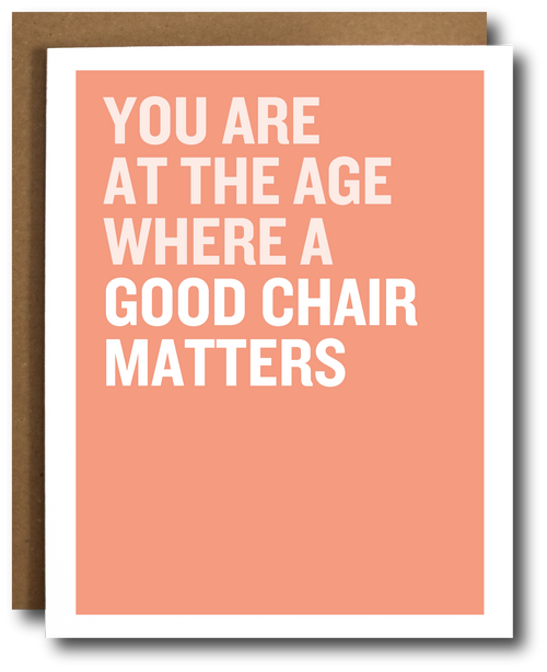 A greeting card with a salmon pink background and bold white text reading 'YOU ARE AT THE AGE WHERE A GOOD CHAIR MATTERS', shown with a brown kraft envelope behind it on a white background.