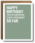 A birthday card with a sage green background and bold white text reading 'HAPPY BIRTHDAY YOU'VE SURVIVED EVERY PRESIDENT SO FAR,' shown with a brown envelope behind it on a white background.