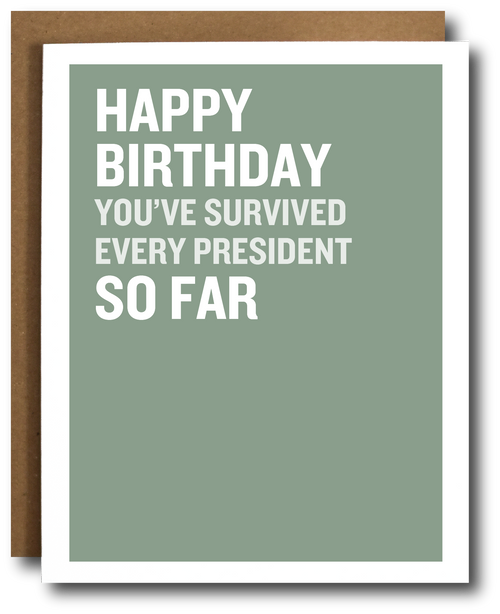 A birthday card with a sage green background and bold white text reading 'HAPPY BIRTHDAY YOU'VE SURVIVED EVERY PRESIDENT SO FAR,' shown with a brown envelope behind it on a white background.