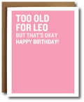 A pink greeting card with bold white text reading 'TOO OLD FOR LEO BUT THAT'S OKAY HAPPY BIRTHDAY!' shown with a brown envelope behind it on a white background.