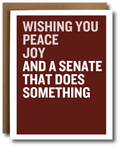 A greeting card with a dark red-brown background and bold white text reading 'WISHING YOU PEACE JOY AND A SENATE THAT DOES SOMETHING,' shown with a brown envelope behind it on a white background.