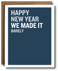 A greeting card with a dark blue front and white border, shown with a brown envelope behind it. The card reads, 'HAPPY NEW YEAR WE MADE IT BARELY' in white uppercase text.