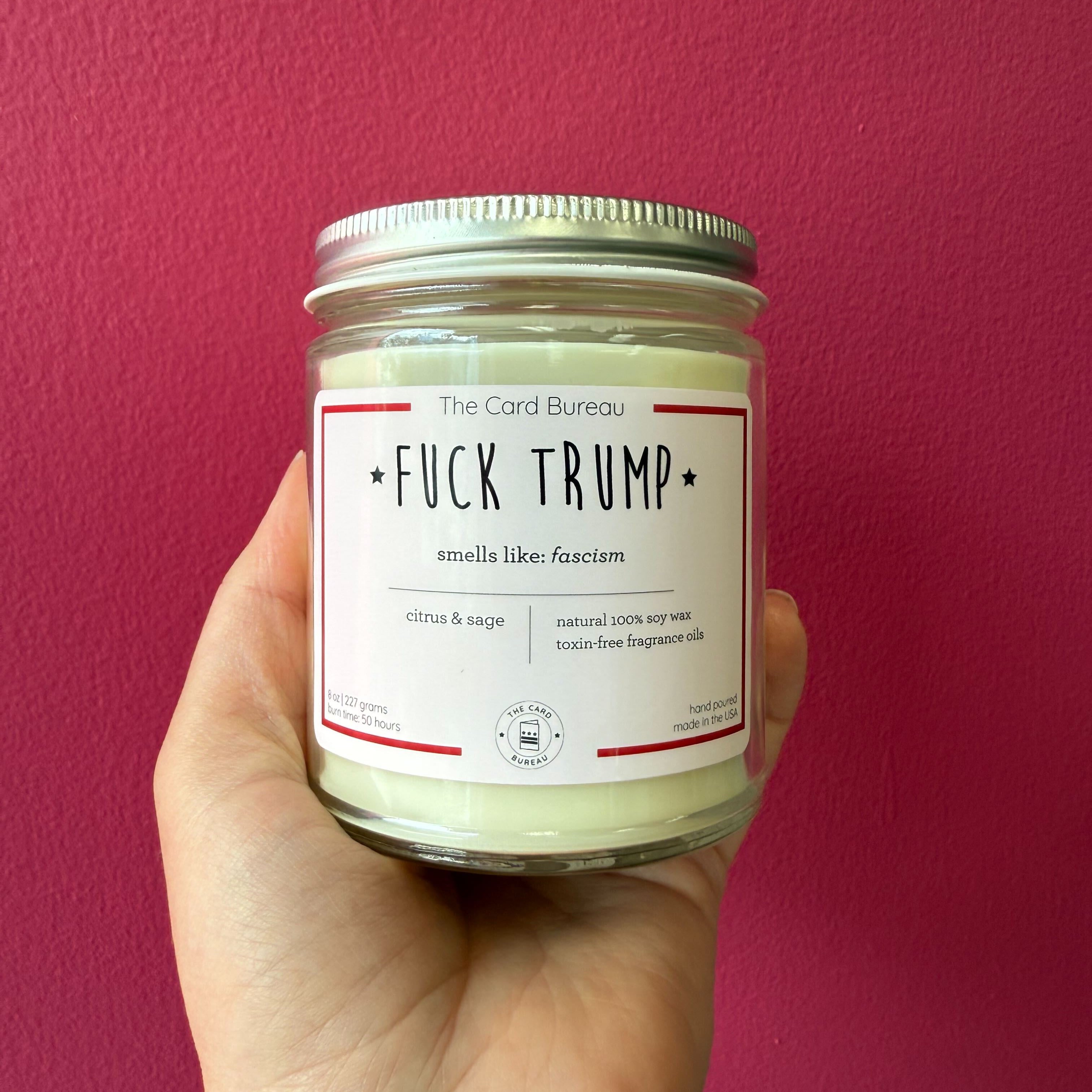 F*ck Trump candle by The Card Bureau — hand-poured soy wax candle scented with citrus and sage, labeled with bold political statement 'smells like: fascism'; 8 oz glass jar with minimalist white and red design, toxin-free fragrance oils, 50-hour burn time, made in the USA